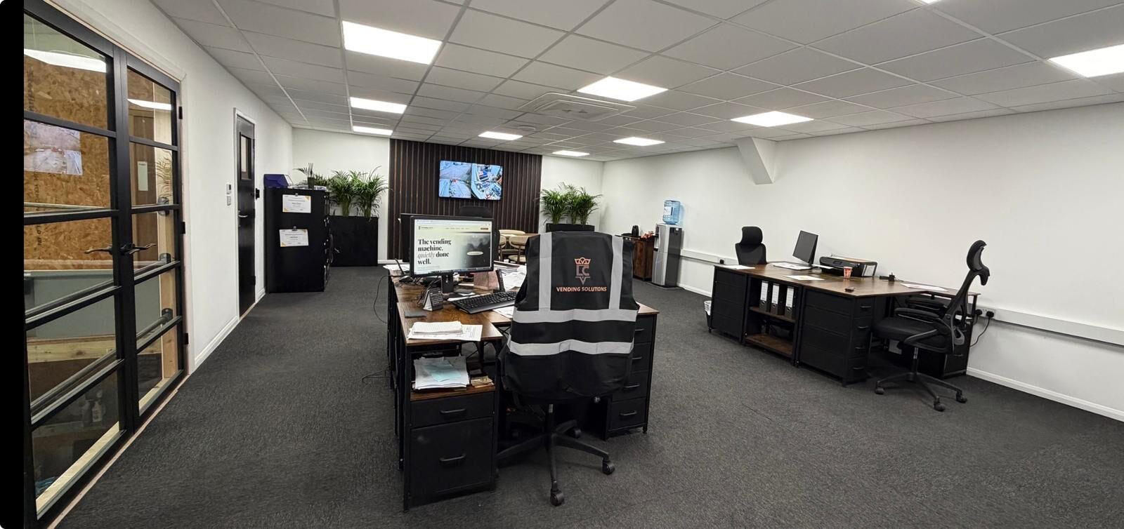 LC Vending Solutions office in Basildon — embroidered hi-vis vest on a chair, multiple workstations, CCTV monitors and the website live on the monitor in shot.